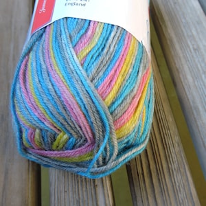 May include: A skein of yarn with a multi-coloured striped pattern in shades of blue, pink, yellow, and grey. The yarn is wrapped around itself and is sitting on a wooden surface.