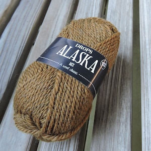 May include: A ball of Drops Alaska Mix yarn in a warm brown hue. The yarn is wrapped with a black label that reads "DROPS ALASKA MIX" and "A wool classic". The label also includes a small circular emblem indicating "100% wool".