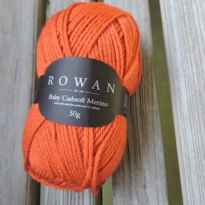 Könnte beinhalten: Ein leuchtend orangefarbener Knäuel Rowan Baby Cashsoft Merino-Garn, 50g, liegt auf einer verwitterten Holzoberfläche. Das schwarze Etikett zeigt den Markennamen und Produktdetails. Das Garn besteht aus extrafeiner Merinowolle und Kaschmir.