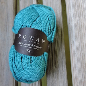 Könnte beinhalten: Ein türkisfarbener Ball Rowan Baby Cashsoft Merino Garn mit einem schwarzen Etikett, auf dem "ROWAN EST 1978 Baby Cashsoft Merino made with extra fine merino wool & cashmere 50g" steht.