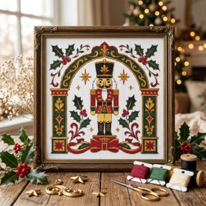 May include: A framed cross-stitch embroidery featuring a central nutcracker figure standing on a red bow, framed by a green and gold archway decorated with holly leaves and red berries, set against a white fabric background with gold stars.