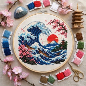 May include: A circular wooden embroidery hoop containing a cross-stitch design of a large blue ocean wave, Mount Fuji, a red sun, and pink cherry blossom branches. The hoop is surrounded by various colored embroidery floss bobbins, a small wooden pagoda model, a ceramic mountain ornament, and gold-colored metal scissors.