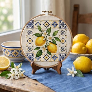 Op de afbeelding: Een ronde houten borduurring met een kruissteekontwerp van twee gele citroenen met groene bladeren en witte bloesems, omlijst door een blauw en geel tegelpatroon op een lichte stoffen achtergrond, geplaatst op een kleine houten standaard.