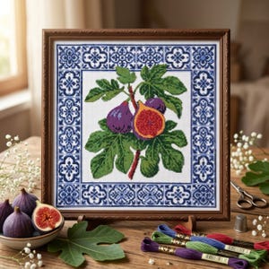 May include: A framed cross-stitch embroidery featuring a central illustration of a fig branch with green leaves and purple figs, surrounded by a blue and white geometric tile-patterned border. The artwork is displayed in a brown wooden frame.