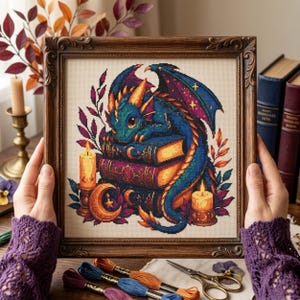 May include: A framed cross-stitch embroidery featuring a teal dragon resting on a stack of books, surrounded by two lit candles, a crescent moon, and foliage. The artwork is displayed in a dark brown ornate wooden frame with a textured fabric background.