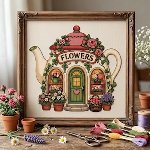 以下が含まれることがあります： FLOWERSという文字が正面に書かれた、ティーポット型の家を描いた額装済みのクロスステッチ刺繍。緑のドア、ピンクと赤のバラ、鉢植え、つる植物がクリーム色の布地にデザインされています。