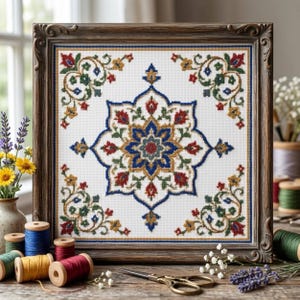 May include: A square cross-stitch embroidery featuring a central blue, red, and gold star-shaped floral motif surrounded by four matching corner floral patterns, all encased in a dark brown ornate wooden frame with a visible textured fabric base.