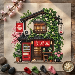 May include: A cross-stitch embroidery on beige fabric depicting a two-story Japanese noodle shop with green vines, a red vending machine, and red banners with the text Udon. Pink cherry blossoms and a bowl of white noodles are visible nearby.