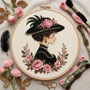 May include: A cross-stitch embroidery project in a circular wooden hoop featuring a profile portrait of a person wearing a black hat adorned with pink roses and feathers, surrounded by a floral wreath of pink roses on a cream-coloured fabric.
