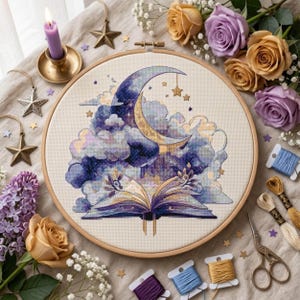 May include: A circular wooden embroidery hoop containing a cross-stitch design of an open book with purple and blue clouds emerging from its pages, topped by a crescent moon and stars, set against a cream-colored fabric background.