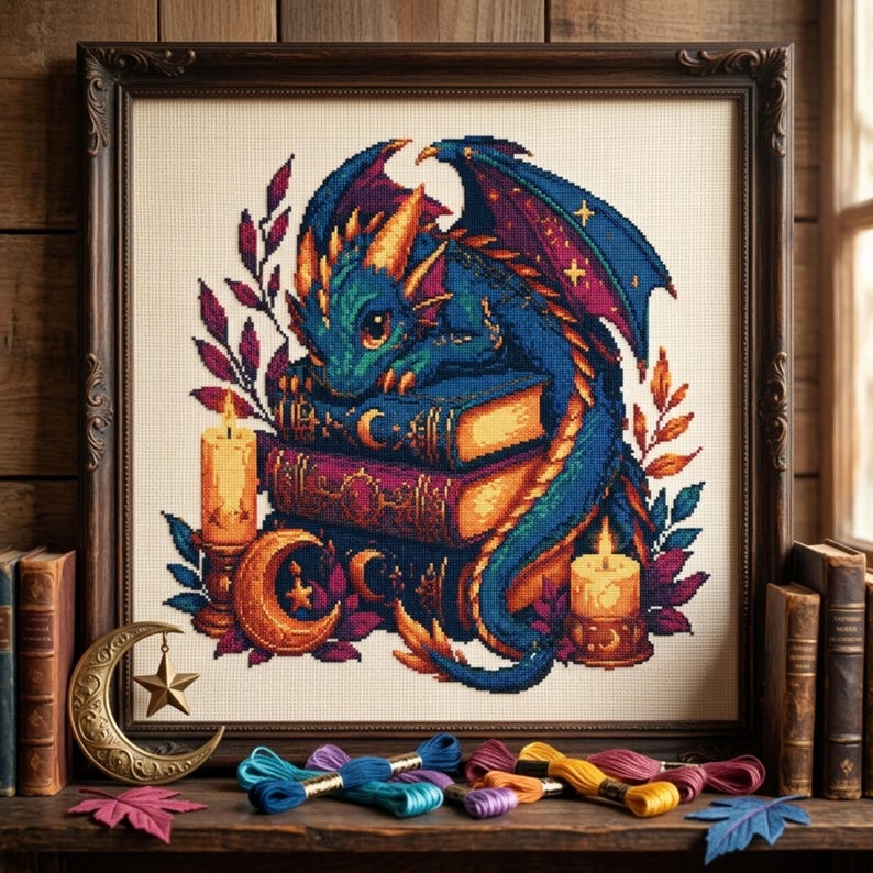 May include: A framed cross-stitch embroidery featuring a blue dragon resting on a stack of books, surrounded by two lit candles, crescent moon motifs, and foliage. The artwork is displayed with colorful embroidery floss skeins and leaves.
