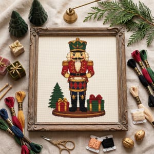 Könnte beinhalten: Eine gerahmte Kreuzstichstickerei, die einen Nussknacker-Soldaten in einer rot-blau-goldenen Uniform beim Trommeln zeigt. Das Motiv umfasst einen kleinen grünen Baum und zwei verpackte Geschenke auf einem braunen Sockel in einem Holzrahmen.
