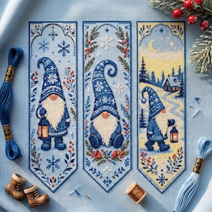 May include: Three vertical cross-stitch patterns on light blue fabric featuring gnomes in blue hats with snowflake motifs. The designs include botanical borders, lanterns, and a winter landscape with a cabin. Blue embroidery floss and small boots are visible.