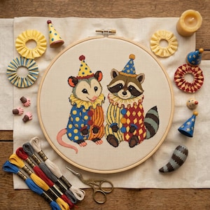 May include: A circular embroidery hoop containing a cross-stitch pattern of an opossum and a raccoon dressed as clowns in colourful polka-dot and diamond-patterned outfits with ruffled collars and party hats, surrounded by craft supplies and accessories.