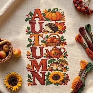 Könnte beinhalten: Eine Kreuzstichstickerei auf cremefarbenem Stoff mit dem Wort AUTUMN in großen orangefarbenen Buchstaben. Das Design zeigt einen Kürbis, einen Igel, einen Korb mit Äpfeln, eine Teekanne, Pilze und eine Sonnenblume, umgeben von bunten Herbstblättern.