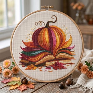 May include: A cross-stitch embroidery project in a round wooden hoop featuring a pumpkin resting on an open book. The design uses vibrant orange, yellow, red, and green threads on cream-colored fabric, with a curly stem and scattered autumn leaves.