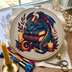 May include: A cross-stitch embroidery in a circular wooden hoop featuring a blue and green dragon resting on a stack of books. The design includes two lit candles, a crescent moon, and foliage in shades of purple and orange on beige fabric.