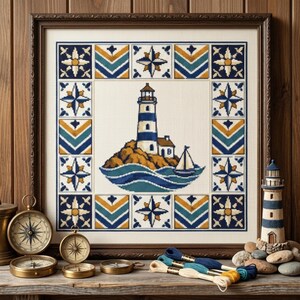 Op de afbeelding: Een ingelijst kruissteek borduurwerk met een centrale vuurtoren op een rotsachtig eiland met een zeilboot, omringd door een decoratieve rand van geometrische patronen en kompassterren in blauw, geel en groenblauw op witte stof.