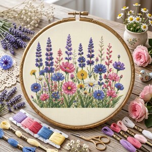 May include: A circular wooden embroidery hoop containing a cross-stitch pattern of a wildflower meadow. The design features tall purple lupines, pink cosmos, yellow daisies, and blue cornflowers on a cream-colored fabric background.