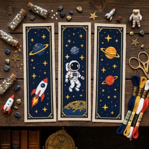 Op de afbeelding: Drie rechthoekige kruissteek-bladwijzers met ruimtethema's op een donkerblauwe achtergrond. De ontwerpen bevatten een raket, planeten met ringen, een astronaut en sterren, omrand door een crèmekleurige stoffen rand met gouden stiksels.