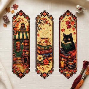 May include: Three vertical cross-stitch bookmarks on white fabric. The left shows a shop front with autumn leaves. The center features a steaming teacup on a stack of books. The right depicts a black cat reading a book with a candle and mug.