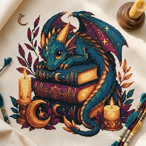 Puede incluir: Un patrón de punto de cruz sobre tela color crema que muestra un dragón verde azulado con cuernos dorados descansando sobre una pila de tres libros. El diseño incluye dos velas encendidas en candelabros, un dije de luna creciente y follaje.