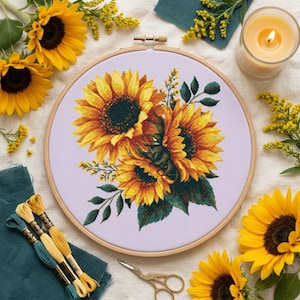 May include: A circular wooden embroidery hoop containing a cross-stitch pattern of three vibrant yellow sunflowers with dark brown centers and green leaves, set against a light lavender fabric background, surrounded by yellow floral accents.