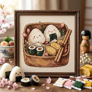 May include: A framed cross-stitch embroidery depicting a bento box filled with two smiling onigiri, two sushi rolls, and two tamagoyaki egg rolls, accented with small pink cherry blossom petals and a pair of wooden chopsticks tied with string.