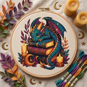 May include: A circular wooden embroidery hoop containing a cross-stitch pattern of a teal and gold dragon resting on a stack of books, surrounded by candles and foliage. The design features vibrant colors, including purple and orange accents.