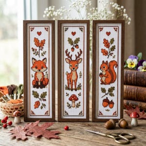 Peut inclure: Trois broderies au point de croix encadrées verticalement sur le thème de la forêt. À gauche, un renard, au centre, un cerf avec un champignon, et à droite, un écureuil avec des glands. Chaque pièce présente des feuilles de chêne et des cœurs.