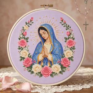 Könnte beinhalten: Ein runder Stickrahmen mit einem Kreuzstich-Porträt der Jungfrau Maria, die einen blauen Mantel und ein cremefarbenes Gewand trägt. Sie ist von einem Kranz aus rosa und weißen Rosen auf einem hellvioletten Stoffhintergrund umgeben.