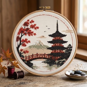May include: A circular wooden embroidery hoop containing a cross-stitch design of a Japanese landscape. The scene features a multi-tiered pagoda, a red arched bridge, a snow-capped mountain, and a tree with red leaves against a white fabric background.