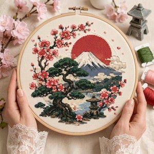 以下が含まれることがあります： 木製の円形刺繍枠に、雪を頂いた山、大きな赤い太陽、濃い緑色の松の木、ピンク色の桜の枝がクロスステッチで描かれています。木の根元には小さな石灯籠が配置されています。