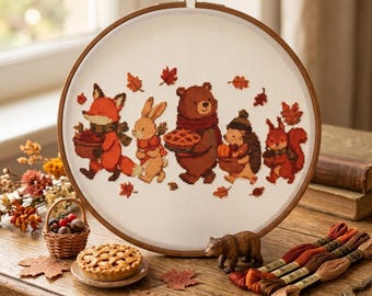 May include: A round wooden embroidery hoop containing a cross-stitch pattern of a fox, rabbit, bear, hedgehog, and squirrel walking together. The animals are depicted in autumn colors with falling leaves, a pie, and pumpkins on a white fabric background.
