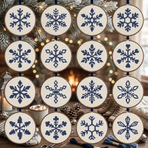 May include: A set of sixteen circular embroidery hoops arranged in four vertical columns, each containing a unique dark blue cross-stitch snowflake pattern on a cream-colored fabric background, suspended by dark blue ribbons.