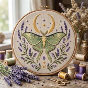 May include: A circular wooden embroidery hoop containing a cross-stitch design of a pale green luna moth centered beneath a yellow crescent moon. The moth is framed by sprigs of purple lavender, with small yellow and purple star accents.