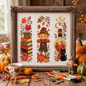 May include: A framed cross-stitch embroidery featuring three vertical panels with autumn themes: a stack of books with a mug, a scarecrow in a hat, and a potion bottle with a black cat, all surrounded by orange pumpkins and embroidery floss.