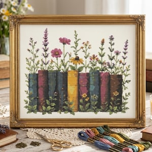 May include: A framed cross-stitch embroidery featuring a row of colorful book spines with various wildflowers growing out of them. The books are in shades of blue, yellow, and pink, while the flowers include purple lavender and yellow daisies.