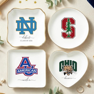 May include: Four white ceramic trinket dishes with gold rims. Each dish features a different college logo: Notre Dame, Stanford, American, and Ohio. The Notre Dame dish has the name "Hobart" and "CLASS OF 2026".