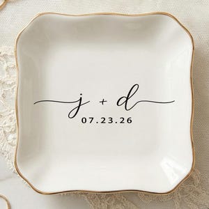 Puede incluir: Un plato de cerámica blanco con borde dorado, con las iniciales "j + d" y la fecha "07.23.26" en escritura negra. El plato está rodeado de anillos de oro y pendientes de perlas, ideal para una boda o compromiso.