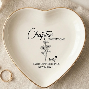Op de afbeelding: Een hartvormig wit keramieken schaaltje met een goudkleurige rand. Het midden bevat zwarte tekst met Chapter TWENTY-ONE, een bloemenillustratie, de naam Emily met een klein hartje en de zin EVERY CHAPTER BRINGS NEW GROWTH.