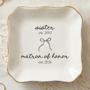 May include: A white ceramic trinket dish with a gold rim, featuring the text "sister est. 2002" and "matron of honor est. 2026" with a bow graphic. The dish is surrounded by jewelry, including rings and earrings.