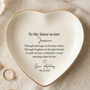 Peut inclure: Un vide-poche en céramique blanche en forme de cœur avec un bord doré. Le centre porte le texte imprimé en noir : To My Sister-in-law, Jessica, un poème sur l'amitié, un cœur, Love, Ashley, et la date 08.22.2026. Plusieurs bagues dorées l'entourent.