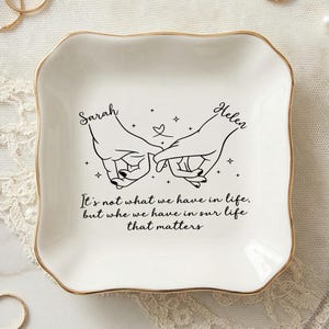 Può includere: Un vassoio portagioie in ceramica bianca con bordo dorato, con un disegno a tratto nero di due mani che si tengono per il mignolo. I nomi "Sarah" e "Helen" sono sopra le mani, con la citazione "Non è quello che abbiamo nella vita, ma chi abbiamo nella nostra vita che conta."