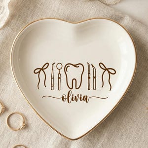 Op de afbeelding: Hartvormige witte keramische sieradenschaal met een gouden rand, met tandheelkundige instrumenten en een tandafbeelding. De naam "olivia" staat eronder geschreven. Verschillende gouden ringen liggen rond de schaal.