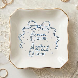 May include: A white ceramic trinket dish with a gold rim, featuring blue illustrations and text. The design includes a bow, baby bottle, champagne bottle, and the words "mom EST. 1999" and "mother of the bride EST. 2026".
