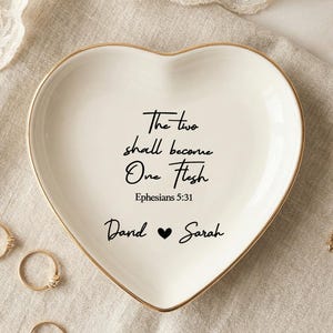May include: A heart-shaped white ceramic ring dish with a gold-painted rim. The center features black text reading "The two shall become One Flesh Ephesians 5:31" followed by "Dand ♥ Sarah". Five gold-toned metal rings are arranged around the dish.