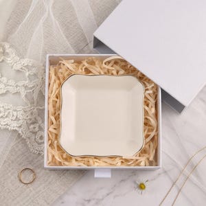 May include: A square, cream-colored ceramic trinket dish with gently scalloped edges and a thin gold-toned rim. The dish is nestled inside a white gift box filled with light-colored shredded paper filler, presenting a clean and simple aesthetic.