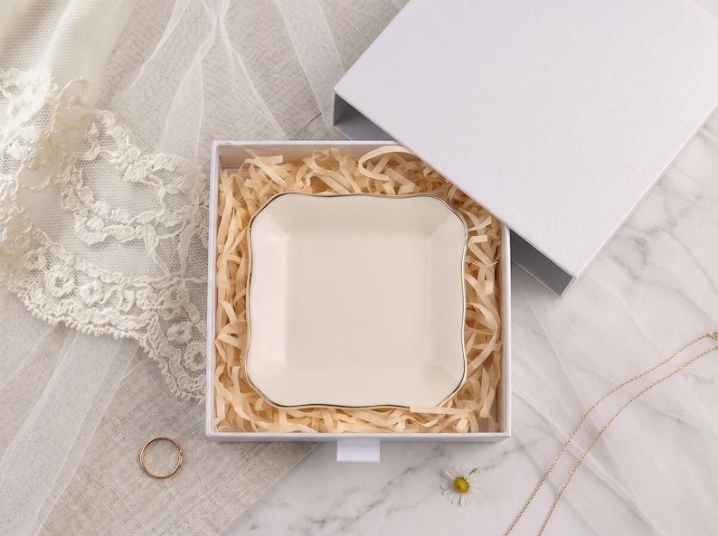 May include: A square, cream-colored ceramic trinket dish with gently scalloped edges and a thin gold-toned rim. The dish is nestled inside a white gift box filled with light-colored shredded paper filler, presenting a clean and simple aesthetic.