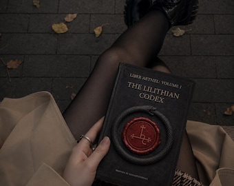 May include: A black hardback book titled Liber Aethel: Volume I The Lilithian Codex, featuring a circular snake emblem surrounding a red wax seal with a symbol, and the text Written by LunarPathways at the bottom of the front cover.